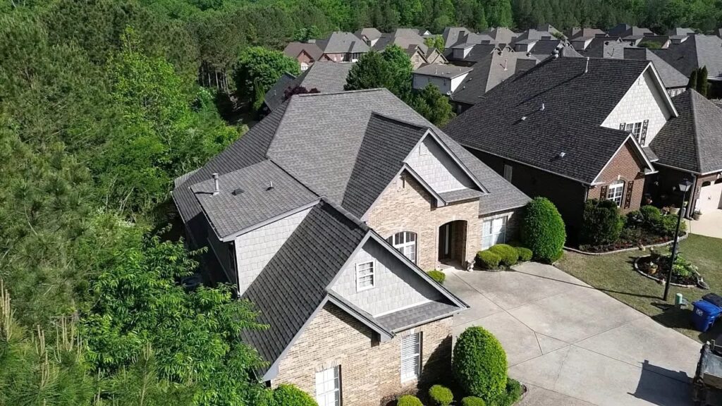 Aerial view of a tan brick home with a gray architectural shingle roof, concrete driveway, and wooded lot beside neighboring