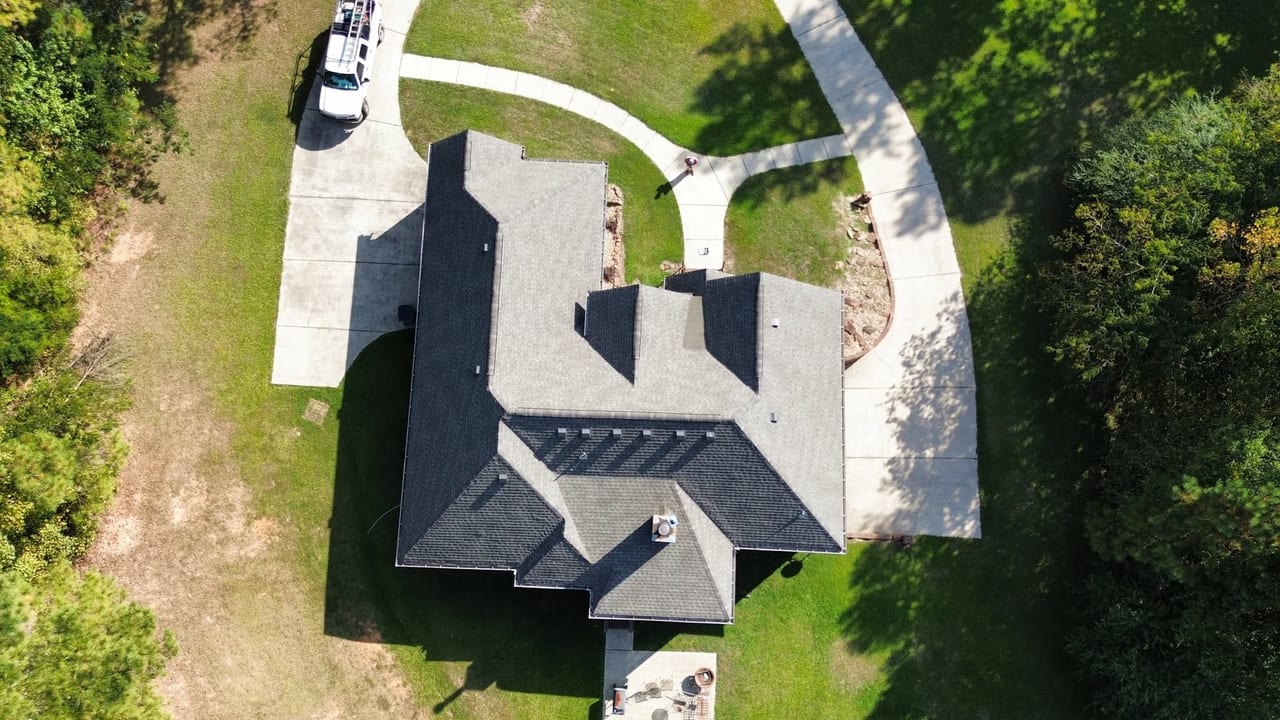Overhead drone view of a house with a new dark gray shingle roof, concrete driveway, and lawn surrounded by trees.