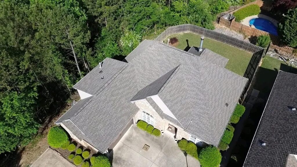 Aerial view of a brick home with a new gray architectural shingle roof, fenced backyard, and wooded lot in Fultondale,
