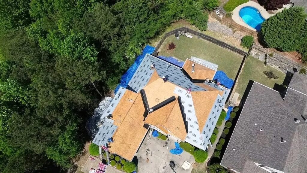 Drone view of a house mid roof replacement with exposed wood decking, synthetic underlayment, and blue tarps around the yard