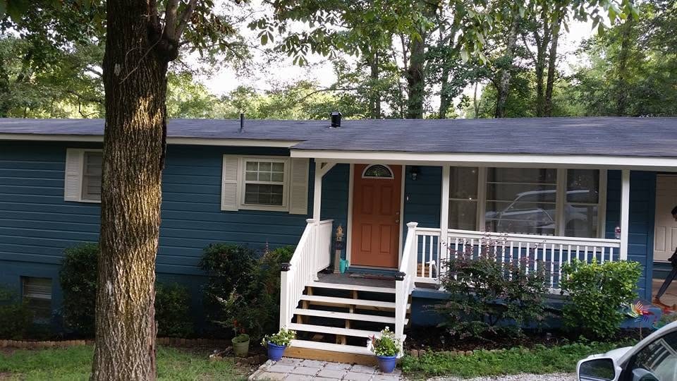 Blue ranch house with dark shingle roof, white porch railing, wood steps, and orange front door shaded by trees