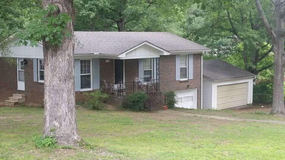 Brick ranch house with gray shingle roof, blue shutters, covered front porch, and attached garage behind a large tree