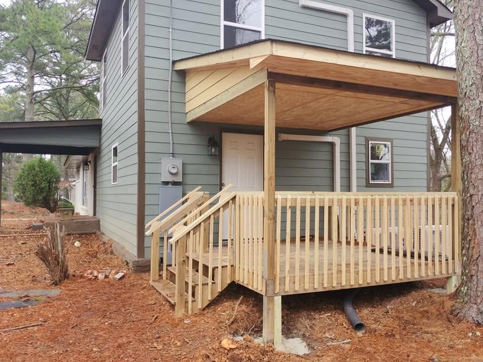 New wooden covered porch with railing and stairs on a two-story gray-sided house among pine trees