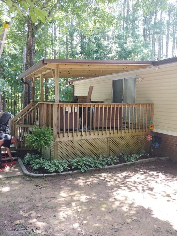Covered wooden deck with railing, lattice skirting, and hosta plants attached to a tan mobile home in a wooded yard.