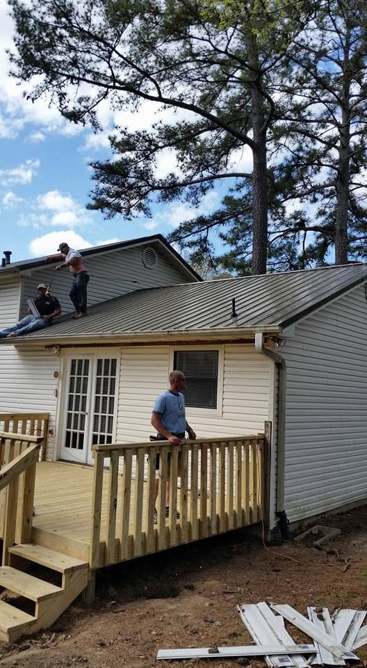 Two workers on a gray metal roof of a white house while another man stands on a new wooden deck below.