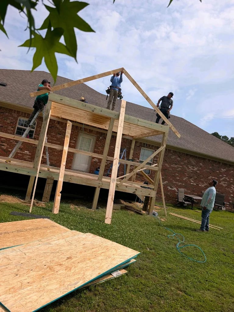 Four workers raising a wood gable rafter over an elevated covered porch addition on a brick home, OSB sheets in foreground