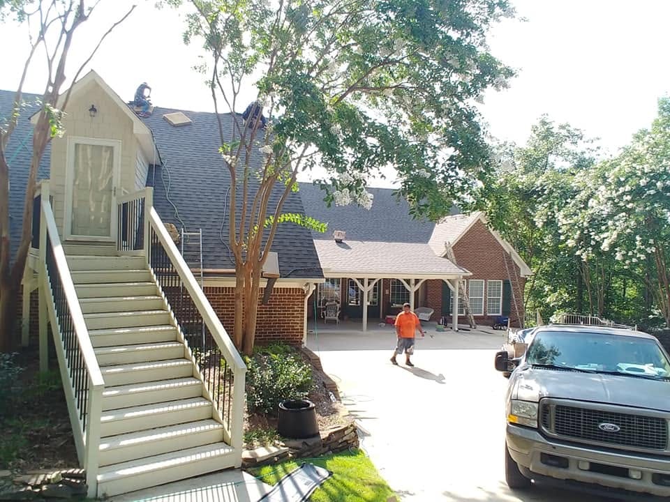 Workers on the shingle roof of a brick home with an exterior staircase and covered patio, truck parked in the driveway.