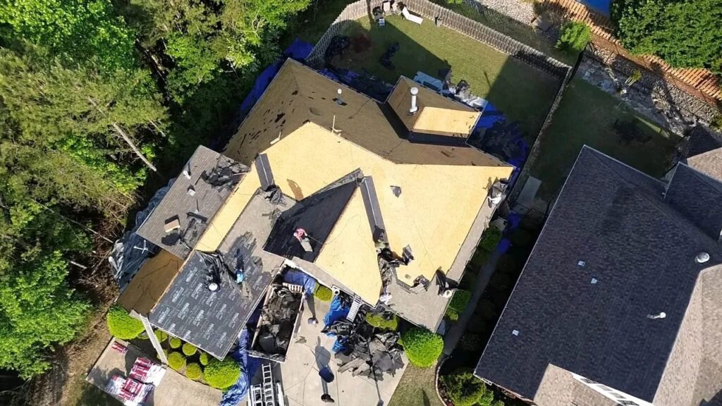 Overhead drone view of a house with shingles removed, bare plywood decking exposed and tarps protecting the yard below.