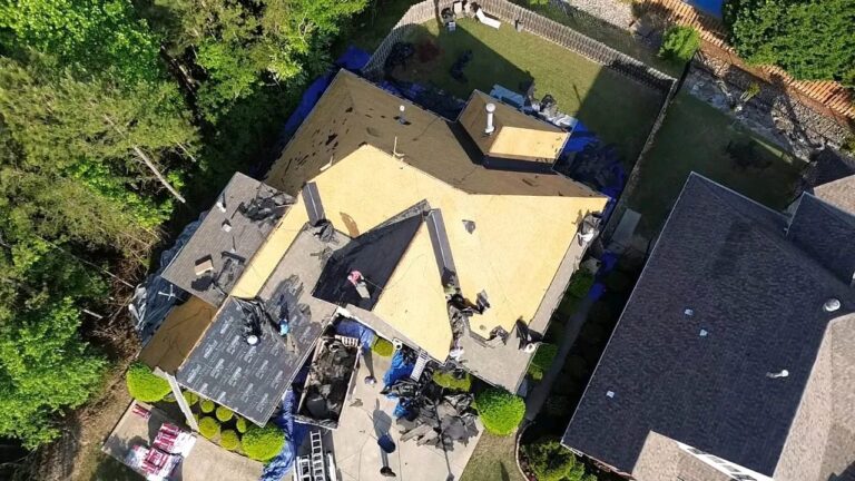Overhead drone view of a house with shingles removed, bare plywood decking exposed and tarps protecting the yard below.