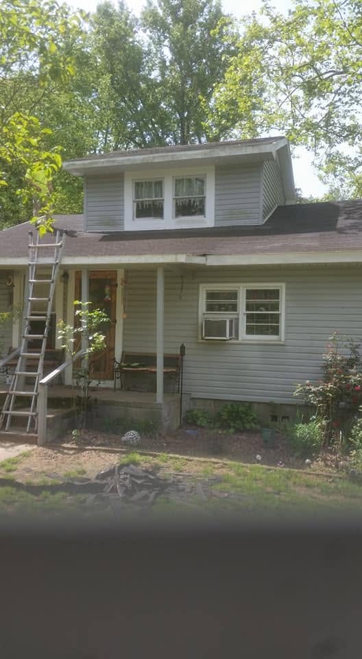 Extension ladder leaning against the porch roof of a gray-sided house with a dormer, set up for roof work.