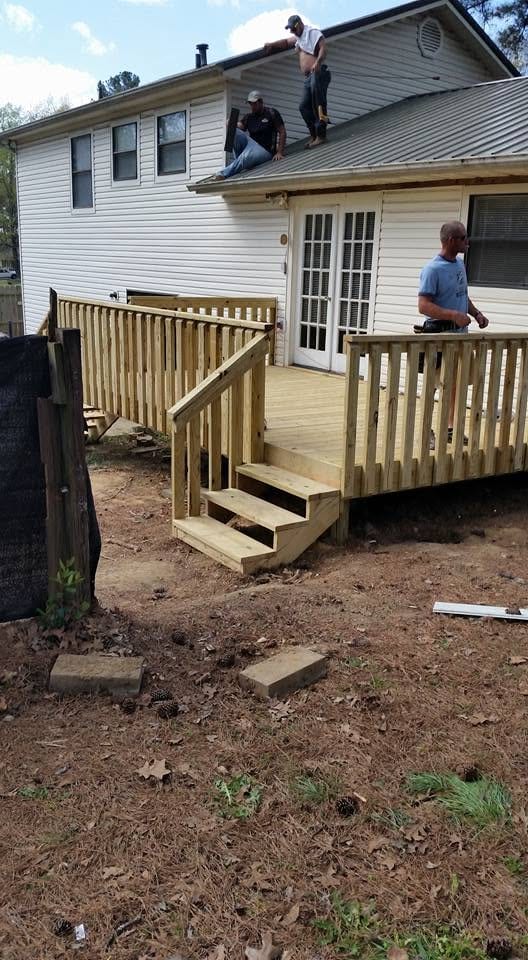 Crew members work on the roof of a white siding home above a newly built pressure-treated wood deck with stairs and railing.
