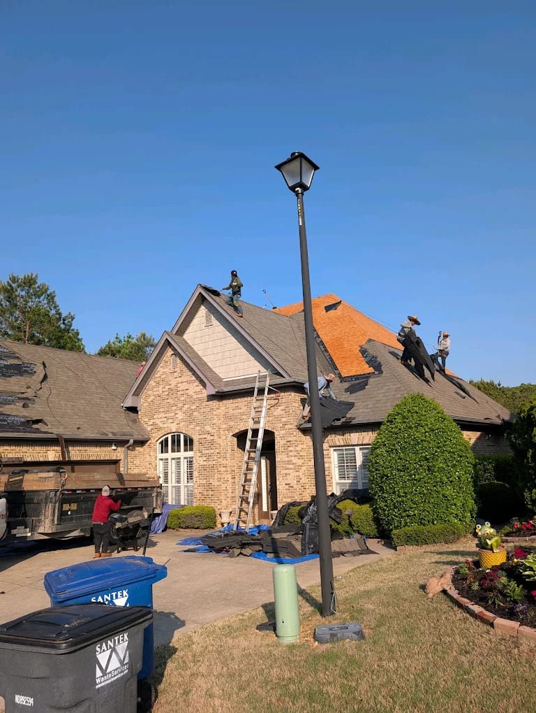 Crew removing shingles from a brick home roof with exposed wood decking, ladder, tarps, and a dump trailer in the driveway