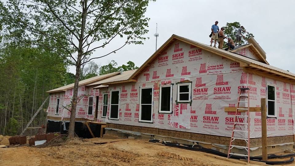 Crew working on the metal roof of a new construction home covered in house wrap, with a ladder at the gable.