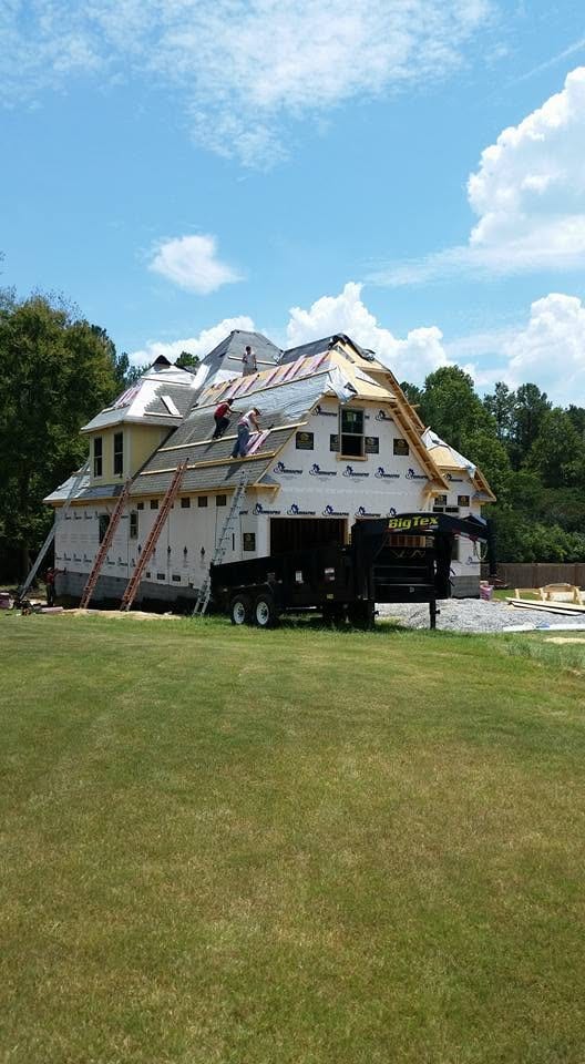Workers on ladders installing shingles on a new construction home with house wrap, black dump trailer parked in front