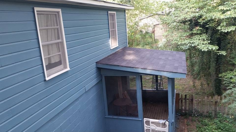 New shingle shed roof over a screened back porch attached to a blue lap-sided house with a wooded backyard.