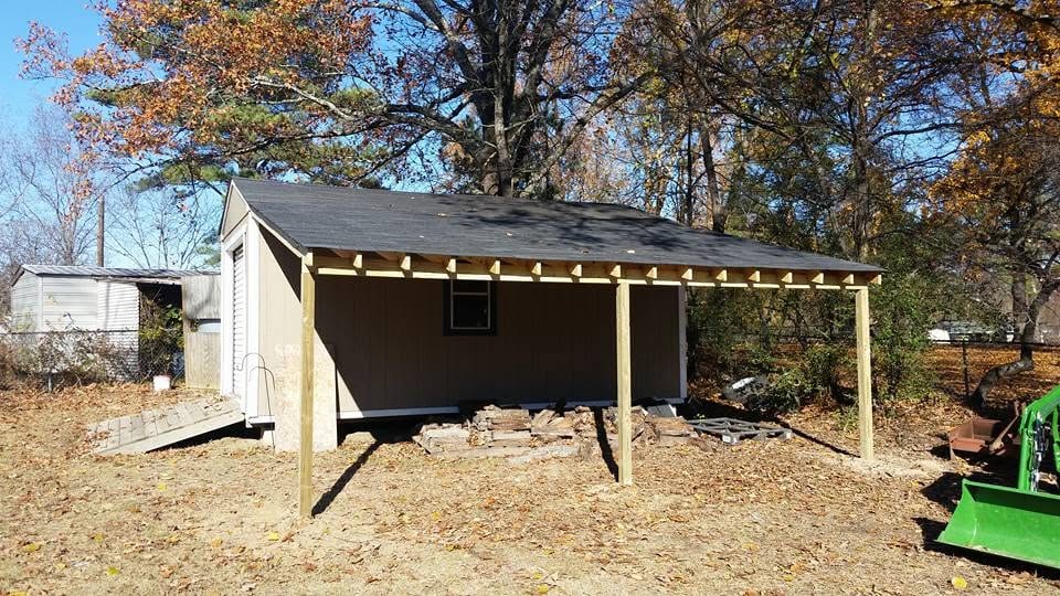 Tan backyard shed with a new lean-to carport addition on wood posts and black roofing, surrounded by fall leaves.