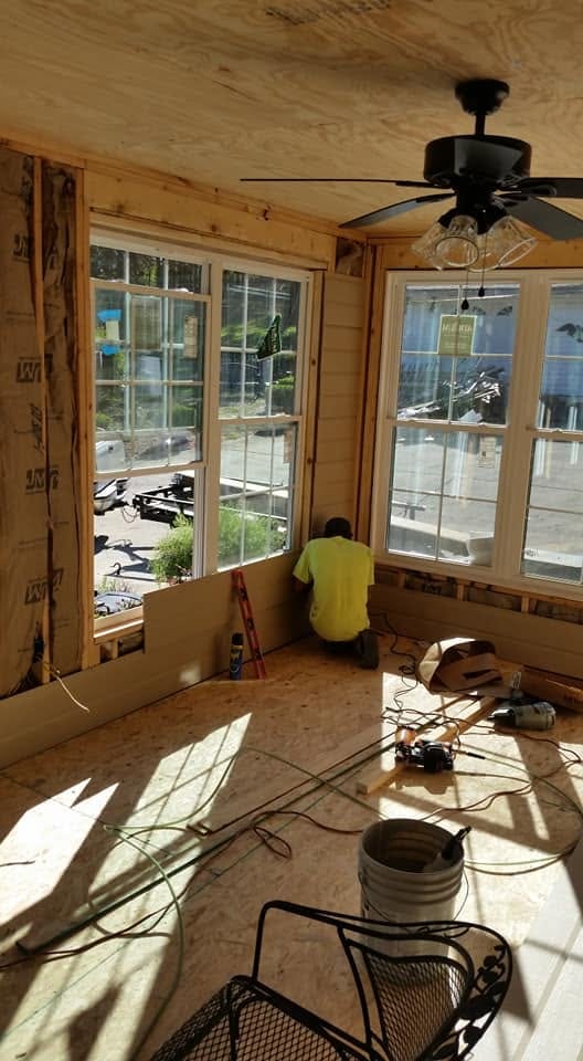 Worker installs wall paneling in a sunroom addition with new double-hung windows, plywood ceiling, and ceiling fan.