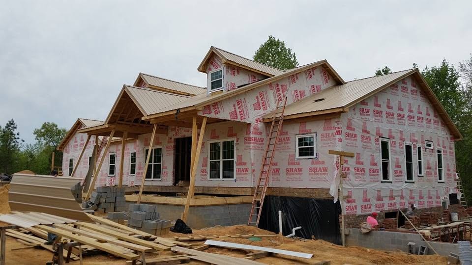House under construction with tan metal roof, printed house wrap, ladder against eave, and lumber stacked in the yard