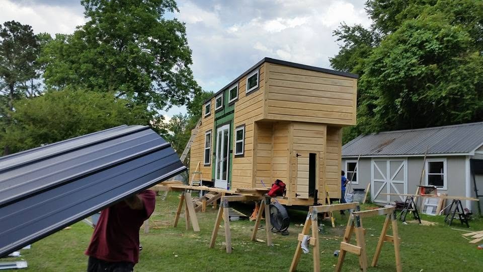Worker carries a black metal roofing panel toward a wood-sided tiny house under construction with sawhorses in the yard.