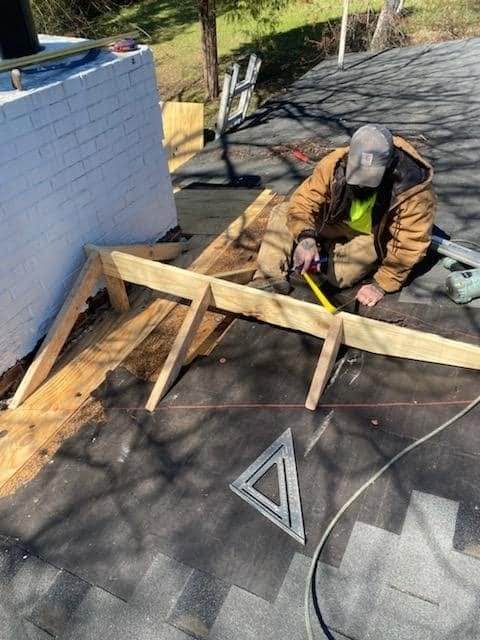 Worker kneeling on a shingle roof measuring new lumber framing for a cricket beside a white brick chimney.